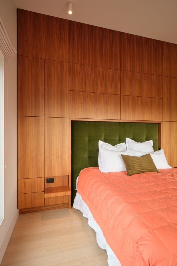 A warm, contemporary bedroom featuring a full-height wall of finely grained timber panelled built-in joinery with discreet seams, integrated bedside shelving, and a dark electrical outlet. The bed has a deep green upholstered tufted headboard recessed into the timber wall, white bedding, and a coral-orange quilt with a textured olive cushion. Pale timber flooring, a white ceiling, and a small ceiling spotlight complete the refined, wood-rich interior.