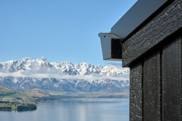Close-up exterior view of a dark, charred timber-clad building with a projecting roof edge, overlooking a calm lake and dramatic snow-capped alpine mountains. The image emphasizes the textured black cladding and strong mountain-lakeside setting.
