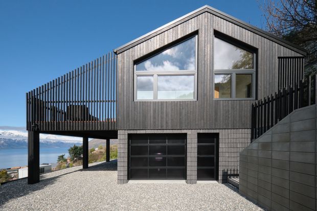 Exterior view of a contemporary elevated home on a sloping site, with a steep gabled roof, dark vertical timber cladding, expansive angular windows, and a lower-level garage. The structure is raised on dark posts, with a screened terrace or balcony extending to the left. A lake and mountains are visible in the background.