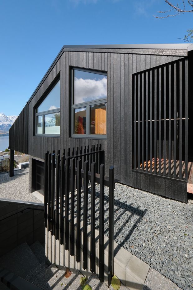 Exterior view of a contemporary dark-clad house on a sloping site. The building features vertical charcoal cladding, large black-framed windows, a low-pitched asymmetrical roofline, and prominent vertical black screening and balustrades around a terrace or entry walkway. Gravel landscaping and stepped concrete paths surround the structure, with glimpses of a mountain and water landscape in the background.