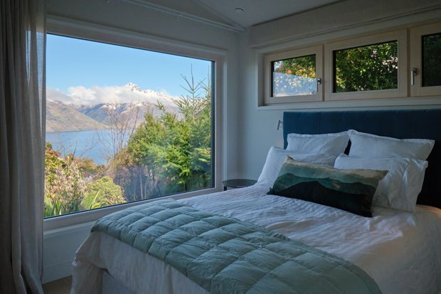 A cozy bedroom with a neatly made bed positioned beside large windows overlooking a blue lake and rugged mountains. The room has white walls and trim, simple horizontal clerestory-style windows above the bed, soft white bedding, a pale blue-green quilt, and a dark teal accent cushion. Natural scenery and daylight dominate the view, creating a calm alpine retreat atmosphere.