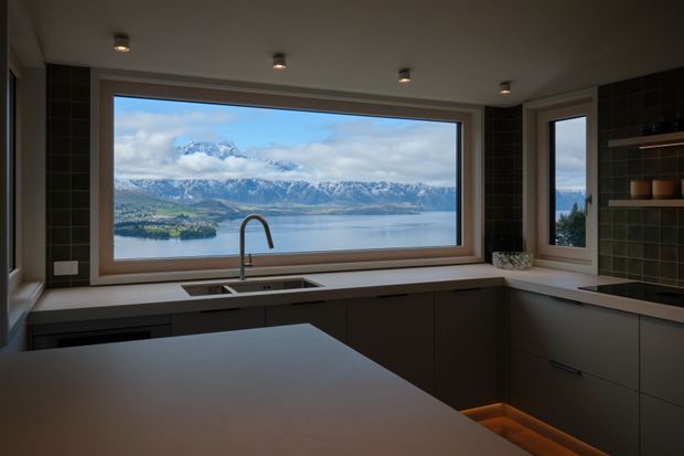 A contemporary kitchen with dark flat-panel cabinetry, a large pale benchtop island, integrated sink and tall curved tap, dark tiled walls, recessed ceiling lights, and expansive timber-framed glazing. The windows provide a prominent view across a lake toward mountainous terrain.
