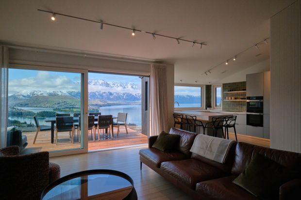 A contemporary open-plan living, dining and kitchen interior with expansive sliding glass doors opening onto a timber deck. The room features a dark leather sofa, glass-topped coffee table, dining table and chairs, a compact kitchen with island bench and integrated appliances, track lighting, timber flooring, and floor-to-ceiling glazing overlooking a lake and mountainous landscape.