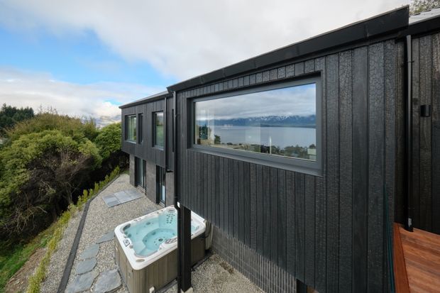 Exterior view of a dark, contemporary multi-level house on a wooded sloping site. The facade is clad in vertically textured dark boards with large aluminium-framed windows, and a covered outdoor spa sits on a gravel and irregular stone-paved terrace. A broad water and mountain landscape is visible through the glazing and beyond the property.