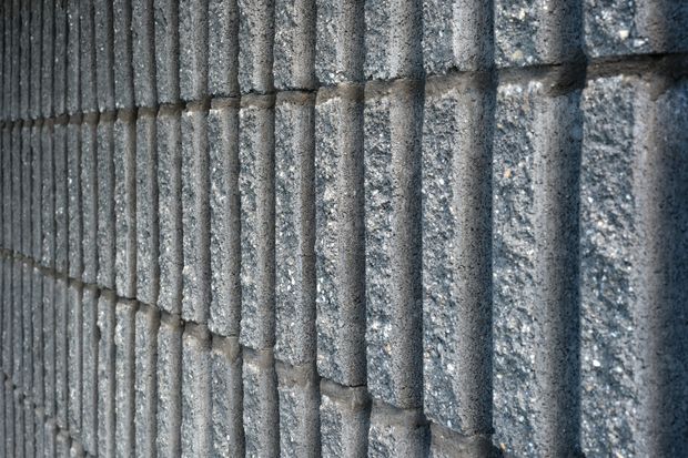 Close-up abstract view of a textured grey masonry wall made from vertically ribbed, rough-faced blocks arranged in staggered courses. The surface has a dark, industrial appearance with strong repetitive vertical lines and subtle tonal variation.