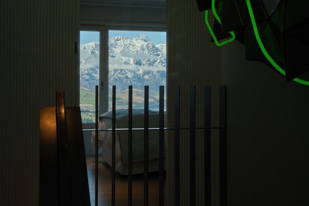 A dark, atmospheric bedroom interior viewed through a partially open vertical-slat partition. A bed is visible beyond the divider, with warm low-level lighting and dark architectural surfaces. Full-height glazing or a door frames a dramatic snow-capped mountain landscape outside. A sculptural dark pendant with green accents hangs in the upper right.