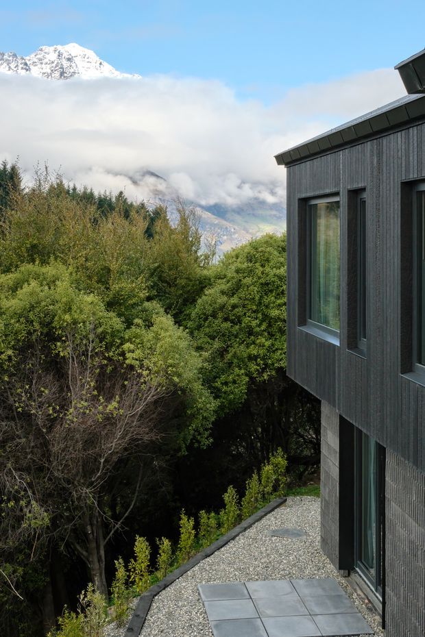 A contemporary dark-clad residential building viewed from an exterior gravel and stone-paved path. Vertical dark timber-like cladding, projecting window frames, and a sloping roof edge define the facade. Dense vegetation surrounds the building, with a snow-capped mountain and cloud-filled valley visible in the background under a clear blue sky.