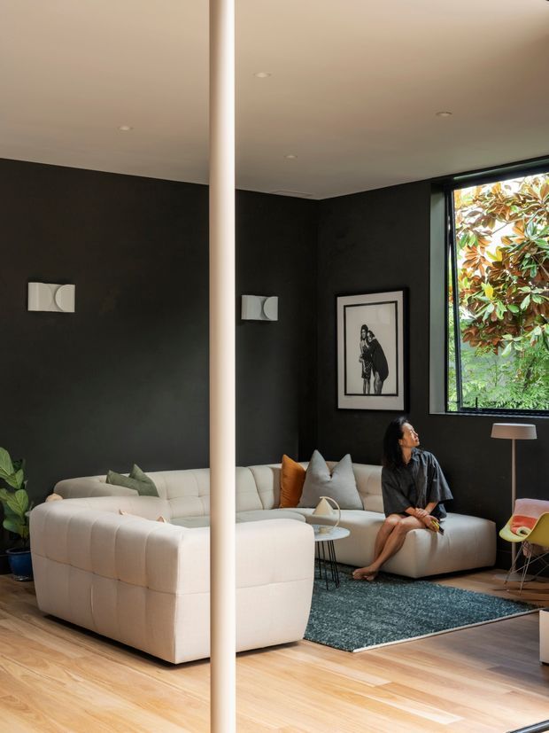A sophisticated living room with charcoal-black walls, a white sculptural sectional sofa, pale timber flooring, and a deep teal rug. A large black-framed window overlooks greenery, while framed artwork, a slender floor lamp, indoor plants, and a small side table complete the calm, high-contrast interior. A prominent vertical structural or decorative column divides the foreground.