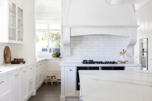 Bright white kitchen with Shaker-style cabinetry, a large marble-look island, white subway-tile splashback, freestanding-style rangehood surround, gas cooktop, and abundant natural light. A windowed breakfast nook and pantry-style cabinetry extend into the background.
