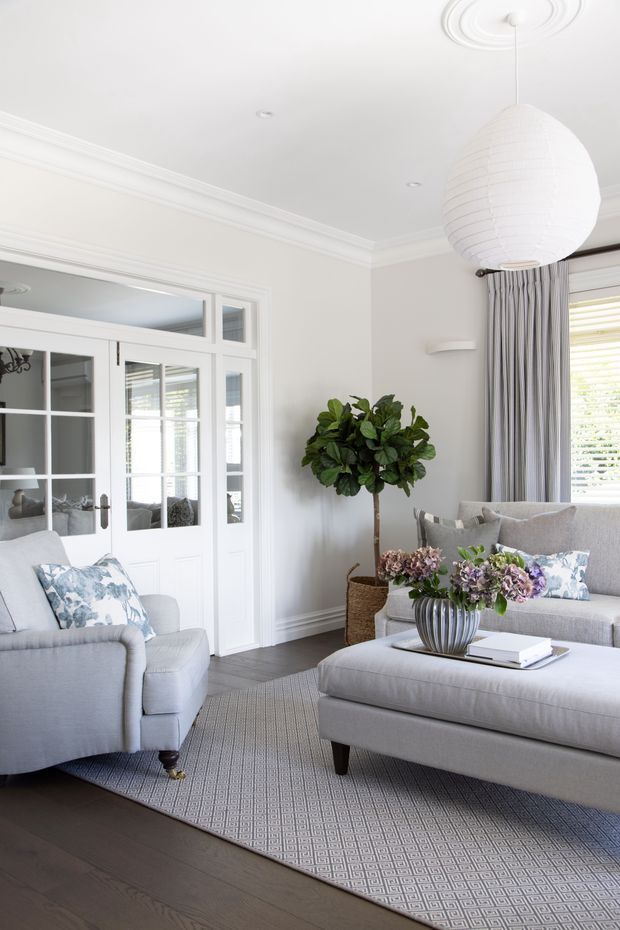 Bright, elegant living room with pale grey upholstered seating, a large matching ottoman, patterned area rug, dark timber flooring, white painted walls and detailed crown moulding. White French doors with glazed panes open toward an adjoining room, while a large spherical pendant light, grey curtains, plantation shutters, decorative cushions, a potted indoor tree and floral arrangement complete the refined interior.