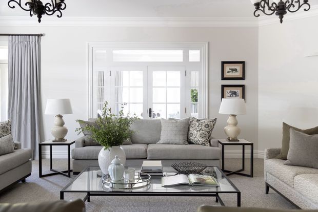 Bright, refined living room with a pale grey sofa and armchairs, glass-top black metal coffee table, matching side tables and table lamps, patterned cushions, framed artwork, and full-height grey curtains. White-trimmed French doors and transom glazing provide abundant natural light, while ornate dark chandeliers add a traditional decorative accent.