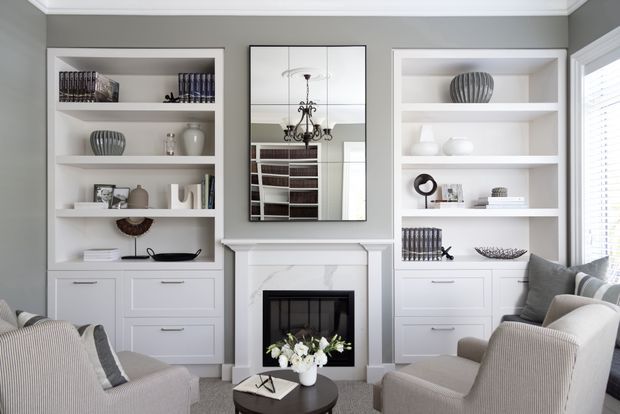 Refined traditional living room with built-in white shelving and drawers flanking a marble-surround fireplace. A large wall mirror reflects the chandelier and additional shelving, while upholstered armchairs, a round dark coffee table, layered books, ceramics, and monochromatic decor create a calm, tailored interior.