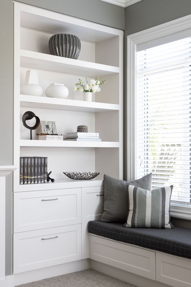 A bright, refined built-in window-seat nook featuring white recessed shelving and lower cabinetry, styled with neutral ceramics, books, and decorative objects. A grey upholstered bench with striped cushions sits beneath a large window with white plantation blinds. The surrounding walls are painted grey, with crisp white trim and soft grey carpet.
