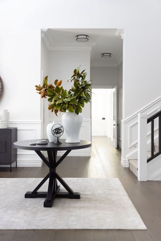 A refined residential entry hall with white paneled walls, decorative ceiling mouldings, pale timber flooring, and a black-framed staircase with light carpeted steps. A round black console table with a sculptural pedestal base sits on a large neutral rug, topped by a white vase with leafy branches and small decorative objects. A bright hallway extends beyond the entry.