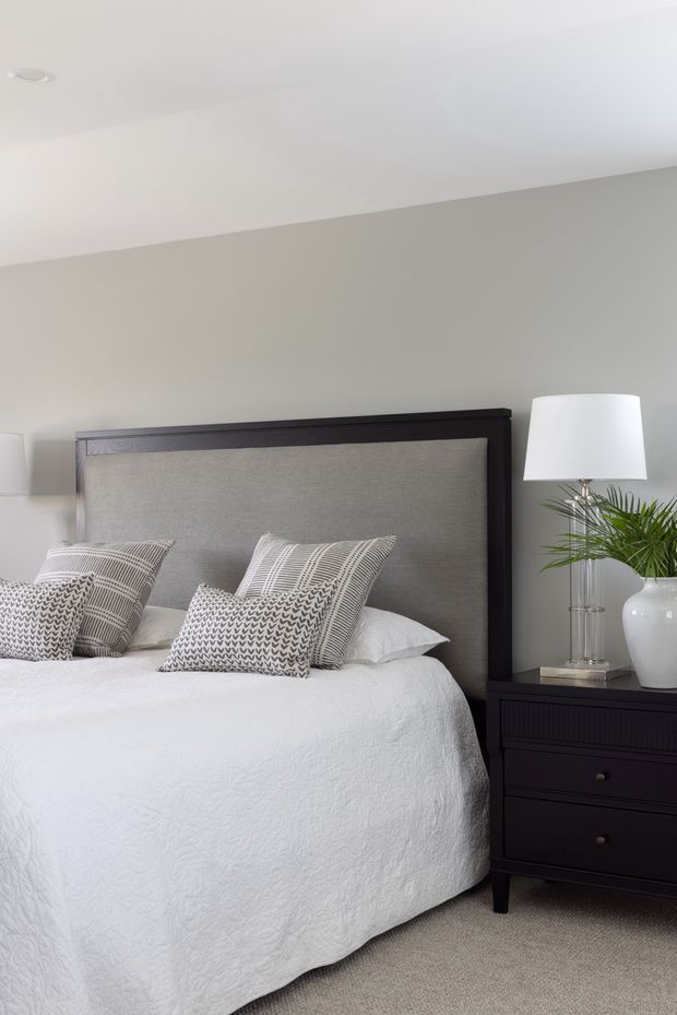 A calm, refined bedroom interior featuring a neatly made bed with a dark timber-framed, upholstered grey headboard, white textured bedding, patterned monochrome cushions, and a matching dark bedside table. A table lamp and potted palm add understated decorative detail. The room has pale grey walls, a white ceiling, and neutral carpet.