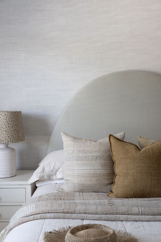 Close-up view of a softly styled bedroom with a large rounded upholstered headboard, layered white and beige bedding, textured neutral cushions, a white bedside table, and a ceramic lamp with a patterned shade. The room has a calm, refined, neutral aesthetic with lightly textured pale wall treatment.