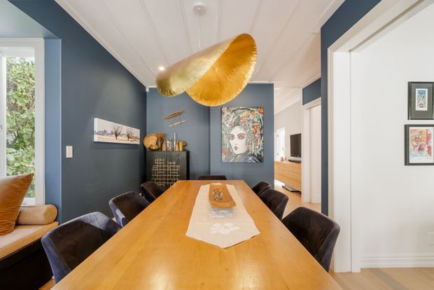 A refined dining room with a long solid timber dining table surrounded by dark upholstered chairs. Deep blue walls contrast with white decorative ceiling panelling and trims, while a large sculptural gold pendant light hangs above the table. Artwork, a dark sideboard, framed prints, and an open doorway toward a timber kitchen area complete the interior.