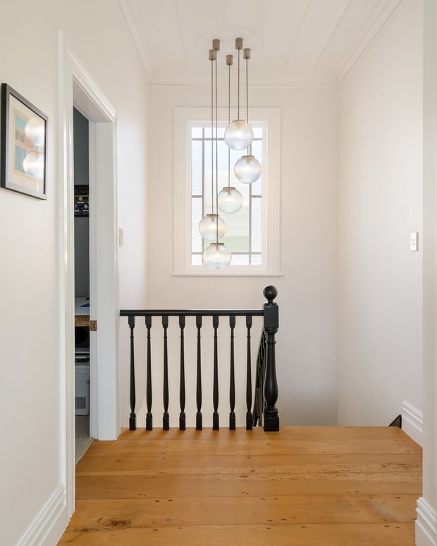 Bright residential stair landing with warm timber floorboards, white walls and decorative trim, a black-painted turned balustrade and newel post, and a vertical cluster of globe pendant lights in front of a tall leaded-style window. A doorway at left opens to an adjacent utility or storage area.