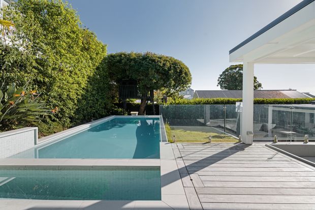 A bright contemporary outdoor entertaining area with a rectangular swimming pool adjoining a broad timber-look deck. The pool is bordered by pale tiled coping and a low tiled retaining wall, with frameless glass balustrades enclosing the deck. A white flat-roofed covered structure with substantial square columns occupies the right side, while mature hedging and landscaped planting provide privacy around the space.