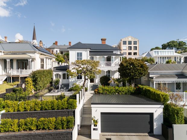 Bright daytime exterior view of a refined residential streetscape, showing closely spaced heritage-style houses with pitched roofs, decorative verandahs, balconies, landscaped front gardens, and a modern integrated garage in the foreground. A church spire and a multi-storey building are visible in the background.