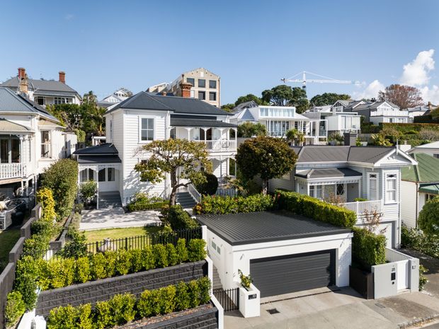 A bright aerial or elevated view of a dense suburban residential neighbourhood, showing several detached and semi-detached homes with pitched metal roofs, weatherboard-style facades, balconies, landscaped gardens, hedges, and a prominent modern double garage in the foreground. The streetscape combines traditional villa-like houses with newer contemporary residences.