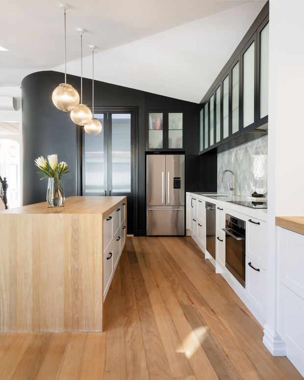 Bright contemporary kitchen featuring a large light-wood island, white shaker-style cabinetry, black walls and upper cabinetry, stainless-steel appliances, a geometric pale tile splashback, timber flooring, and black-framed clerestory-style glazing. Three pendant lights hang above the island, and daylight enters through the extensive upper windows.