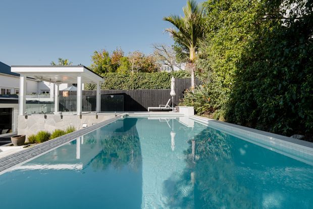 Bright exterior view of a modern residential swimming pool, with the pool dominating the foreground. A glazed pavilion or poolside living area with a flat roof sits to the left, bordered by dark fencing and dense tropical landscaping.