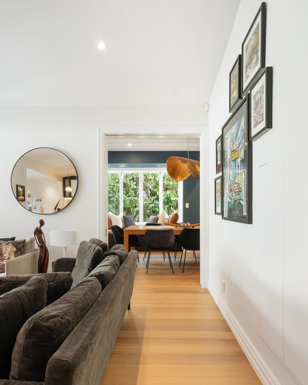 Bright contemporary interior view from a living room toward an adjoining dining area. The foreground contains a large dark grey upholstered sofa, a round wall mirror, artwork, and a floor lamp. Through the wide doorway, a timber dining table with dark upholstered chairs and a sculptural pendant light is visible against a dark accent wall and greenery outside. White walls, recessed lighting, timber flooring, and decorative framed artwork create a refined, warm and inviting space.