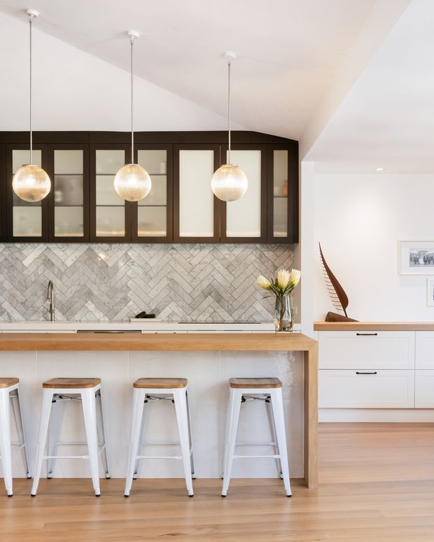 Bright, refined kitchen interior featuring a large timber-topped island with white cabinetry and four white stools, a grey herringbone tile splashback, dark timber-framed glass-front upper cabinets, and three spherical pendant lights. The kitchen opens toward a white built-in storage area with timber countertop and light timber flooring.