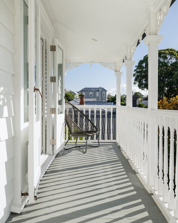 A bright covered front veranda or balcony on a white weatherboard home, featuring ornate decorative fretwork, square columns, turned balustrade details, and grey decking. French doors open onto the veranda, with a woven hanging chair and views toward neighbouring houses and greenery. Strong sunlight casts striped shadows across the floor.