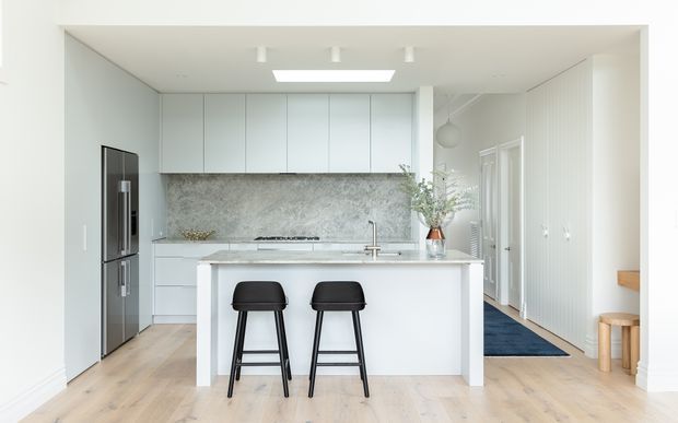 Bright, minimalist contemporary kitchen viewed straight on, featuring a large white island with two black bar stools, pale blue-grey handleless cabinetry, a grey marble-look splashback, integrated stainless-steel refrigerator, light timber flooring, and a hallway extending to the right.