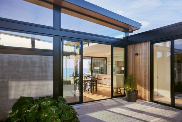 A contemporary single-storey courtyard-facing exterior with extensive black-framed glazing and a partially covered timber-lined roof canopy. Through the glass, a bright open-plan kitchen and dining area is visible, featuring timber flooring, light cabinetry, pendant lighting and bar seating. The foreground has a timber deck or paved terrace with tropical potted planting, while a rendered boundary wall and glimpses of blue water are visible beyond.