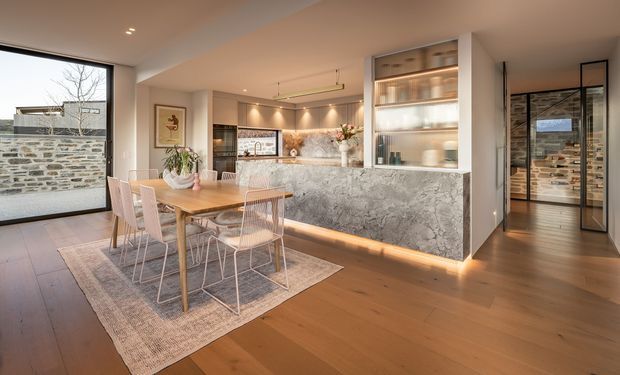Open-plan contemporary kitchen and dining area with warm timber flooring, a timber dining table and pale wire-frame chairs, and a large stone-clad kitchen island. The kitchen features integrated cabinetry, illuminated glass-front display joinery, pendant lighting, recessed spotlights, and soft under-cabinet illumination. Full-height glazing and a glazed hallway door connect the interior to an exterior stone boundary wall and a view toward another room. The palette is warm and neutral, with prominent grey veined stone and light timber.