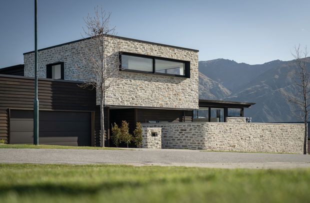 Exterior view of a contemporary two-storey residence with a low, rectilinear form, dark horizontal cladding, extensive black-framed glazing, and light natural stone masonry walls. A garage and enclosed courtyard wall are visible in the foreground, with a dramatic mountain backdrop beyond the property.
