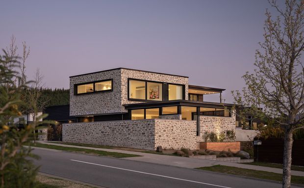 Twilight exterior view of a contemporary multi-level residence, featuring a strong rectangular form, extensive glazing, dark metal framing, and textured light brick masonry walls. The home sits along a suburban street with landscaped planting and a low brick boundary wall.