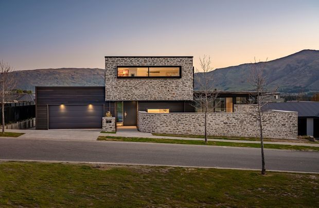 Street-facing exterior view of a contemporary multi-level residence at dusk. The house features a long, low composition with dark metal roofing and cladding, extensive grey irregular-stone masonry across the upper volume and front boundary wall, a recessed entry, horizontal strip windows, and an integrated double garage. Bare deciduous trees and a mountain backdrop frame the property.
