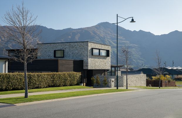Contemporary two-storey residential house in a suburban street, featuring a rectilinear form, dark horizontal cladding, pale textured masonry upper walls, and black-framed windows. The home sits behind clipped hedges and a lawn, with modern street lighting and dramatic mountain ranges in the background.