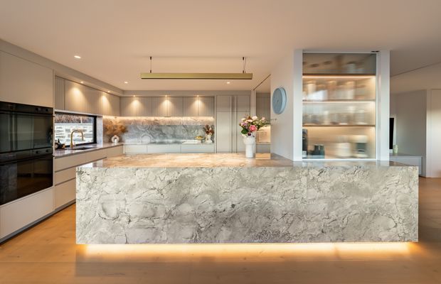 A refined contemporary kitchen viewed front-on, dominated by a large stone-clad island with integrated underlighting. The kitchen features light timber flooring, pale cabinetry, dark integrated appliances, a matching stone splashback, recessed ceiling spotlights, and a suspended linear pendant. A glass-fronted display cabinet and open shelving are visible to the right, with a small dining or living area beyond.