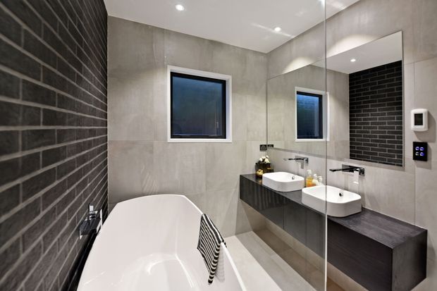 Contemporary bathroom with a freestanding white bathtub, double vessel basins on dark timber-look cabinetry, large mirrored wall, grey stone-look wall and floor tiles, and a dark charcoal brick feature wall. Recessed ceiling lighting and a wall-mounted control panel complete the refined monochromatic scheme.
