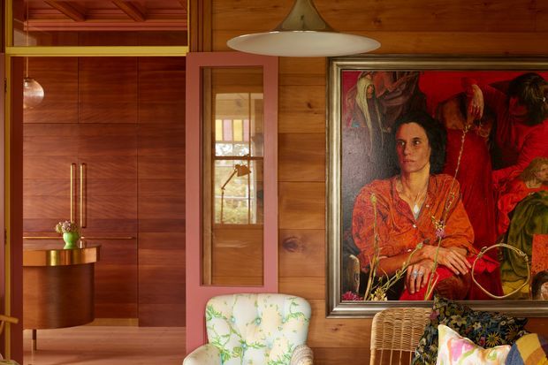 Warm, richly timber-lined interior featuring extensive dark wood built-in cabinetry, a tall glass-fronted door or cabinet, a sculptural pendant light, and an eclectic seating area. A large framed artwork dominates the wall, with woven and floral-upholstered chairs adding colour and texture. The image presents a refined, art-filled living space with strong mid-century character.