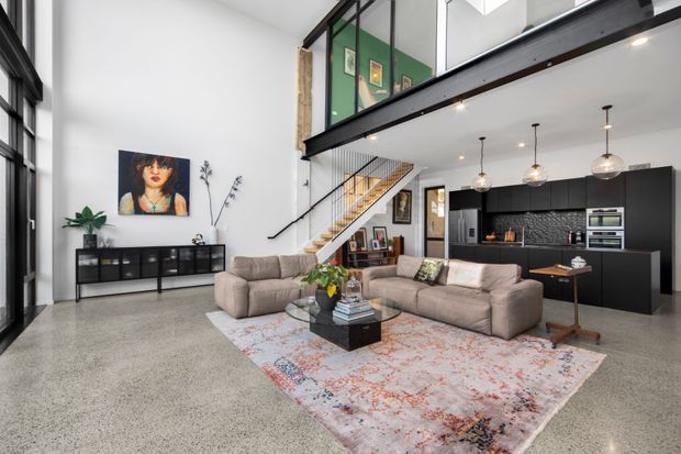 A bright, open-plan industrial-style living space with a double-height ceiling and mezzanine level. The room features polished concrete flooring, a large sectional sofa, a patterned area rug, black-framed glazing, exposed dark steel beams, a floating timber stair, and a compact black kitchen with pendant lighting.