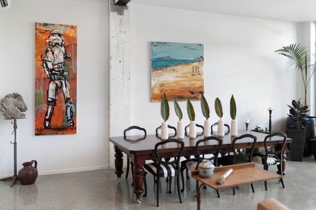 A bright, eclectic dining room with a dark timber dining table and black spindle-back chairs arranged beneath a row of decorative potted plants. White walls, polished terrazzo-style flooring, contemporary paintings, sculptural decor, and large indoor plants create a refined yet artistic interior. A partial living area is visible at the right edge.