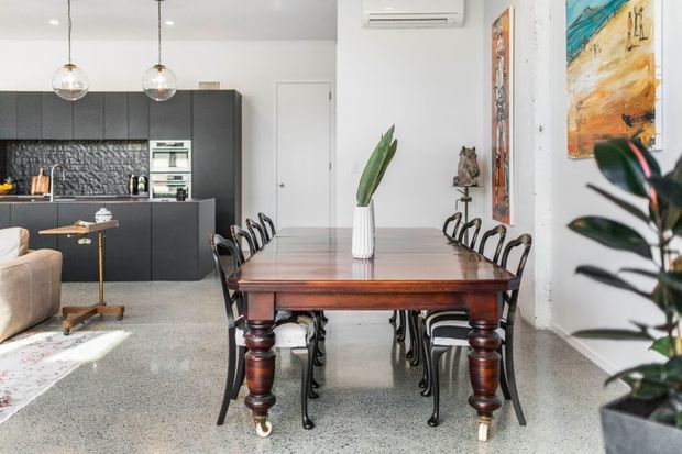 Bright open-plan dining and kitchen interior featuring a substantial timber dining table with turned legs, black metal dining chairs, polished concrete flooring, and a dark contemporary kitchen with a textured splashback. Pendant globe lights, colourful artwork, indoor plants, and white walls add visual interest.