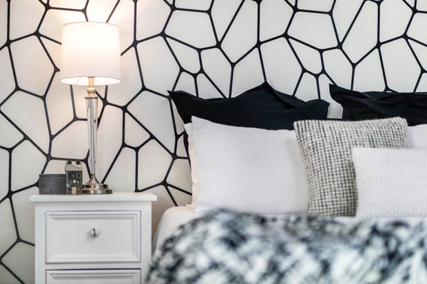 Close-up of a contemporary bedroom vignette featuring a white bedside table with paneled drawers, a chrome table lamp with a white drum shade, layered white, black and textured grey bedding, and a bold black-and-white irregular geometric wall covering behind the bed.
