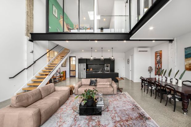 Bright open-plan loft-style interior with a living room, dining area, kitchen, and prominent mezzanine level. The space features exposed concrete and black steel structural elements, polished concrete or terrazzo flooring, white walls, extensive internal glazing, a black kitchen, timber stair treads, and large neutral sofas arranged around a patterned rug and coffee table. A dining table with dark chairs occupies the right side, while a colourful artwork wall and indoor plants add accents.