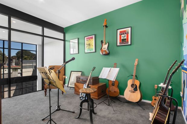 A bright music room or creative studio with a bold green feature wall, several guitars mounted and displayed around the room, a piano, music stands, framed posters, and a glass-panel door opening to an exterior balcony or terrace. The space has grey carpet, black-framed glazing, and abundant natural light.