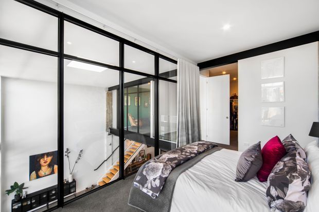 Contemporary bedroom overlooking a stairwell through a black-framed steel-and-glass partition. The room features a white bed with patterned grey bedding and cushions, grey carpet, white walls, recessed lighting, sheer curtains, built-in shelving, and framed wall panels. The glazed partition creates visual connection to the lower level while maintaining separation.