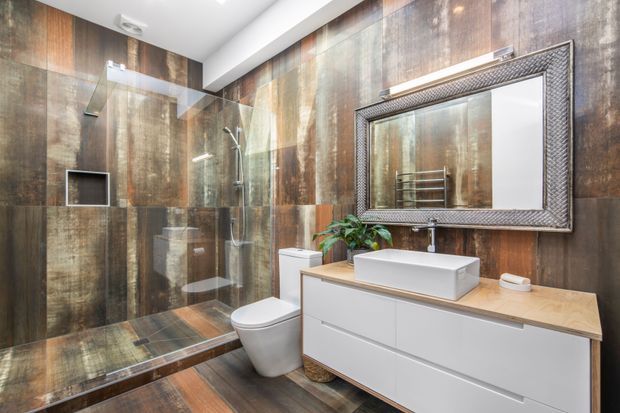 A contemporary bathroom featuring full-height dark timber-look wall and floor tiles, a frameless glass walk-in shower, wall-mounted toilet, and a white vessel basin on a timber-topped floating vanity. A large textured metallic-framed mirror and brushed metal fixtures add contrast, while a small plant provides greenery.