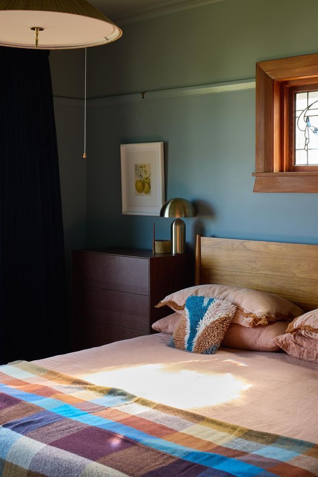 A warmly lit bedroom with deep blue-green painted walls, a timber bedhead, dark timber dresser, brass-toned bedside lamp, framed artwork, and a colourful geometric blanket layered over soft pink bedding. A timber-framed stained-glass window and dark curtain add character, while sunlight creates a cozy, inviting atmosphere.