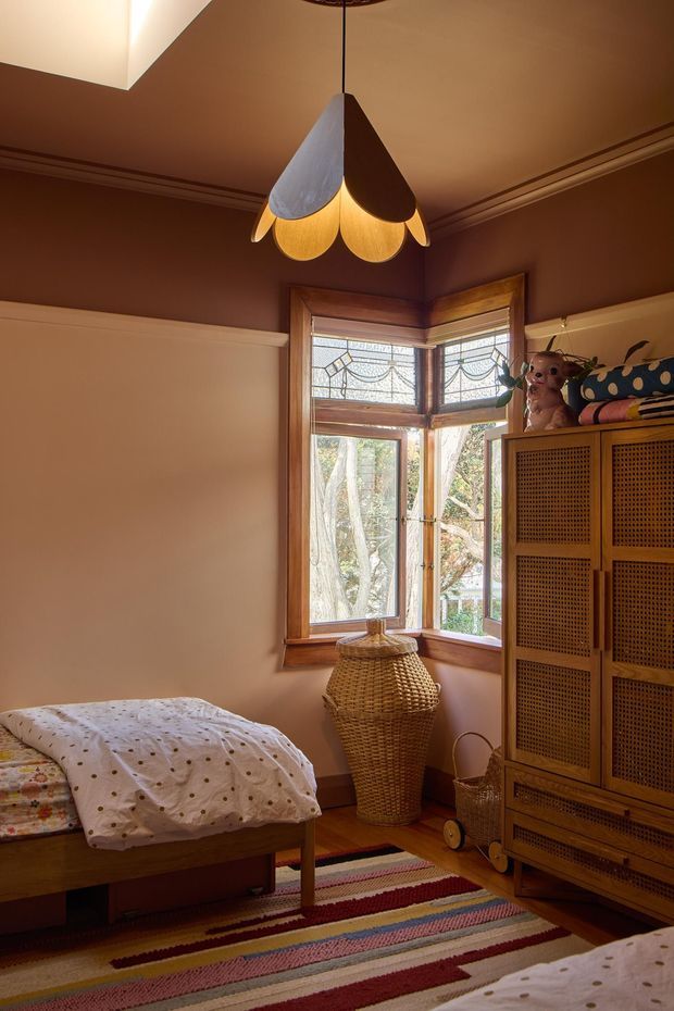 A warm, eclectic bedroom with rich brown walls, timber-trimmed bay windows featuring decorative leadlight glazing, and a distinctive petal-shaped pendant light. The room contains a wooden bed with patterned bedding, a woven rattan wardrobe, a tall wicker basket, a striped woven rug, and playful soft furnishings. Natural light enters through the windows, creating a cosy, characterful atmosphere.